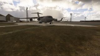 A gray four-engine military transport sits on an apron near hangars and floodlight towers beneath a cloudy sky.