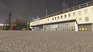 An airport terminal has a glass-fronted departure entrance beside a hangar, floodlight towers, and a parked truck under dark clouds.