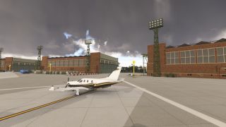 A white single-engine propeller aircraft sits on an apron beside brick hangars and tall floodlight towers beneath dark clouds.