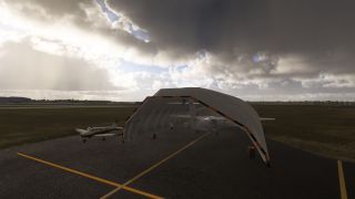 Two light aircraft sit beside and beneath an arched shelter on a yellow-lined apron under dark clouds.