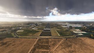 A runway stretches toward the horizon beneath dark clouds, with approach lights, taxiways, and hangars surrounded by dry grass.