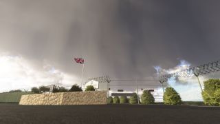 A Union Jack flies above a stone wall beside a gated airfield entrance with barbed-wire fencing beneath dark clouds.