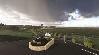 Britannia Gate at RAF Brize Norton stands beside security fencing, with a Union flag, green hangars, and dark clouds overhead.