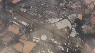 An overhead view shows a single-runway airfield with connecting taxiways, parked aircraft, hangars, and surrounding farmland and residential neighborhoods.