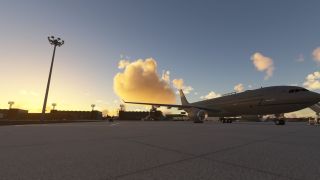 A gray twin-engine airliner sits on an airport apron beside hangars and floodlight towers beneath golden clouds.