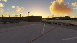 A passenger jet sits on a marked apron beside terminal buildings and floodlight towers beneath golden clouds.