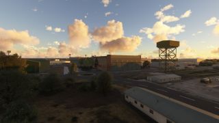 Airport hangars and low service buildings surround a tall lattice-supported tower beneath golden clouds, with a tanker truck parked nearby.