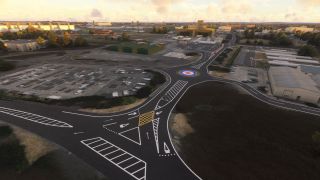 A roundabout with a red, white, and blue roundel connects marked roads beside parking lots, green hangars, and industrial buildings.