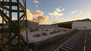 A four-engine military transport aircraft sits outside hangars among maintenance lifts and crates, behind a perimeter fence under golden clouds.