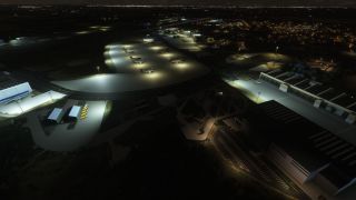 An airport at night features parked aircraft on floodlit aprons, large hangars, blue taxiway lights, and illuminated neighborhoods beyond.