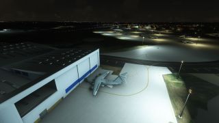 A four-engine turboprop transport sits beside a large hangar at night, with floodlit aprons and other parked aircraft beyond.