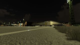 Floodlight towers illuminate an airport apron at night, with parked airliners beside terminal buildings and a bus near the grass verge.