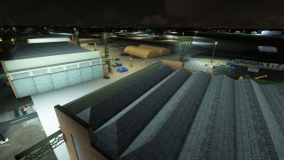 Floodlight towers illuminate airport hangars, parked aircraft, tan shelters, and cargo containers at night, with large corrugated roofs dominating the foreground.