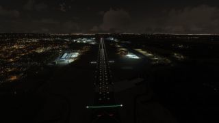 An illuminated runway extends through a nighttime airport, flanked by blue taxiway lights, floodlit buildings, and surrounding city lights beneath clouds.