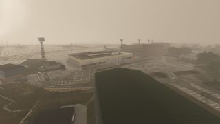 An airport terminal stands beside marked parking areas and floodlight towers, with parked aircraft barely visible through dense haze.