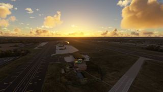 An airfield stretches beneath golden clouds, with a control tower, parked aircraft, hangars, and intersecting paved runways visible from above.