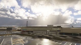 An airport terminal overlooks wet, marked parking bays, with parked aircraft visible behind the building beneath sunlit clouds.