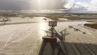 A large jet and several propeller aircraft sit on a wet airport apron beside hangars beneath broken clouds and sunlight.