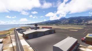 A coastal airport features large hangars, a control tower, and small aircraft on the apron, with populated green hills beyond.