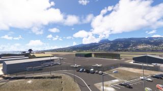 Small aircraft sit beside hangars on a fenced airport apron, with parked cars in front and green mountains beyond.