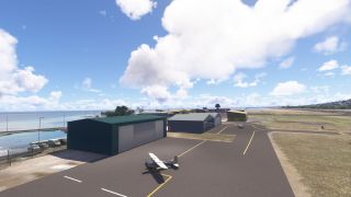 A small high-wing aircraft sits on a coastal airport apron beside hangars, with yellow taxiway markings and a runway beyond.