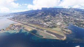 A coastal airport runway extends beside turquoise shallows, with apron buildings and dense hillside development beneath cloud-covered mountains.