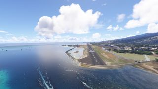 A coastal runway extends beside blue water, with displaced threshold markings, airport buildings, and a populated hillside beneath scattered clouds.