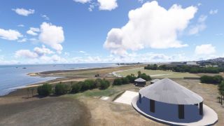 A coastal airport runway extends beside the sea, with conical-roofed structures and scattered palms in the foreground beneath cumulus clouds.