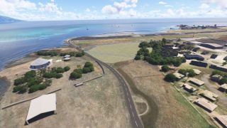 A coastal runway borders turquoise water, with hangars, cylindrical storage tanks, scattered buildings, and a curving road in the foreground.