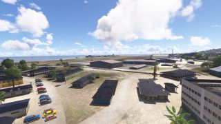 Low-rise airport buildings and parked cars occupy the foreground, with hangars and a coastal runway beneath scattered cumulus clouds.