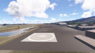 A white circular helipad marks the airport apron beside hangars, with a runway to the left and developed hills beyond.