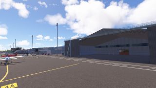 A large open hangar stands beside parked light aircraft, yellow apron markings, and tall floodlights under scattered clouds.