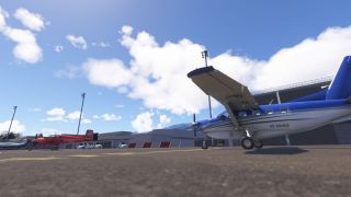 A blue-and-white high-wing turboprop sits on the apron beside hangars, with a red twin-engine aircraft parked farther away.