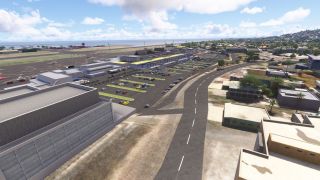 A coastal airport terminal overlooks car parks and a curving access road, with small aircraft parked near taxiways and the sea beyond.
