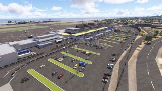A coastal airport terminal overlooks a large parking lot with green medians, while small aircraft sit on the apron beside the runway.