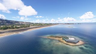 A coastal runway borders blue water, with hillside buildings beyond and a circular antenna structure on a small foreground island.