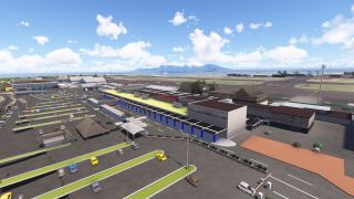 An airport terminal with blue columns faces car-filled parking lots, while taxiways, ocean waters, and a mountainous island stretch behind it.