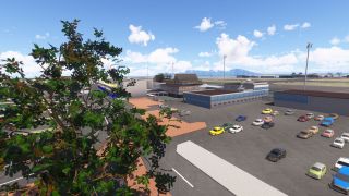 Cars fill a parking area beside airport buildings, with leafy trees in the foreground and mountains beyond the airfield.