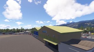 An Air Tahiti Nui hangar with a pale green roof stands beside an apron, with parked cars and forested hills behind.