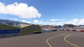 An Air Tahiti Nui hangar borders an apron with curved yellow markings, mobile passenger stairs, and terminal buildings backed by green hills.