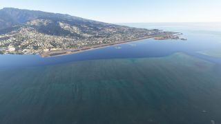 A coastal runway stretches beside blue water, with airport buildings at one end and dense housing climbing the green mountainside.