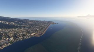 A coastal runway borders a densely built hillside town, with shallow offshore reefs and a mountainous island visible across the water.