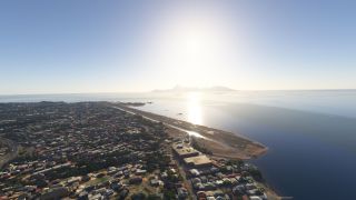 A coastal runway extends beside dense neighborhoods and calm water, with sun glare reflecting toward a mountainous island on the horizon.