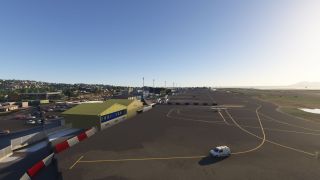 A coastal airport apron features a green hangar, yellow taxiway markings, a small aircraft, and a white service van.