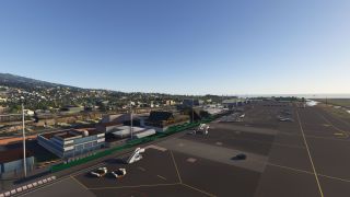 A coastal airport apron contains parked aircraft, mobile stairs, and service vehicles beside terminal buildings, with hillside neighborhoods beyond.