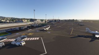 Several business aircraft sit on a marked airport apron beside terminal buildings, with mobile passenger stairs and tall floodlights nearby.