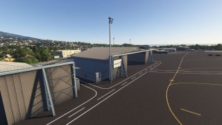 Large gray hangars border an empty apron with yellow taxi lines, white service roads, and tall floodlights beside a hillside neighborhood.