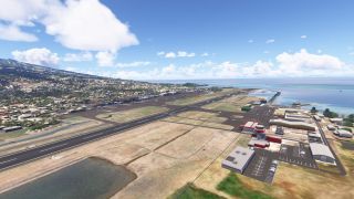 A coastal airport has a long runway, branching taxiways, hangars, and parking areas, with hillside neighborhoods and ocean beyond.