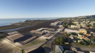 A coastal airport features a runway beside the sea, hangars, a marked helipad, and surrounding hillside neighborhoods.