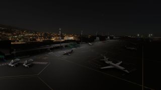 Business jets sit on a floodlit airport apron at night, with mobile boarding stairs, terminal buildings, and hillside lights beyond.